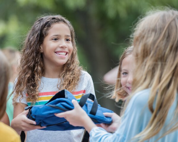 Ein Mädchen übergibt einem anderen Mädchen einen Rucksack und lacht dabei | © Courtney Hale - Getty Images/iStockphoto